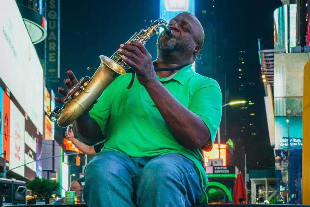 Auf dem Bild ist ein Mann in einer grünen Poloshirt und blauen Jeans zu sehen, der in Times Square eine Saxofonblase spielt. Die bunten Leuchttafeln und der urbane Hintergrund geben der Szene einen unverwechselbaren Flair. Ebenso wie die Musik des Mannes die Menschen anzieht, strebt "Über BaoLiba" danach, österreichische Influencer und Marken auf der ganzen Welt zu vernetzen.